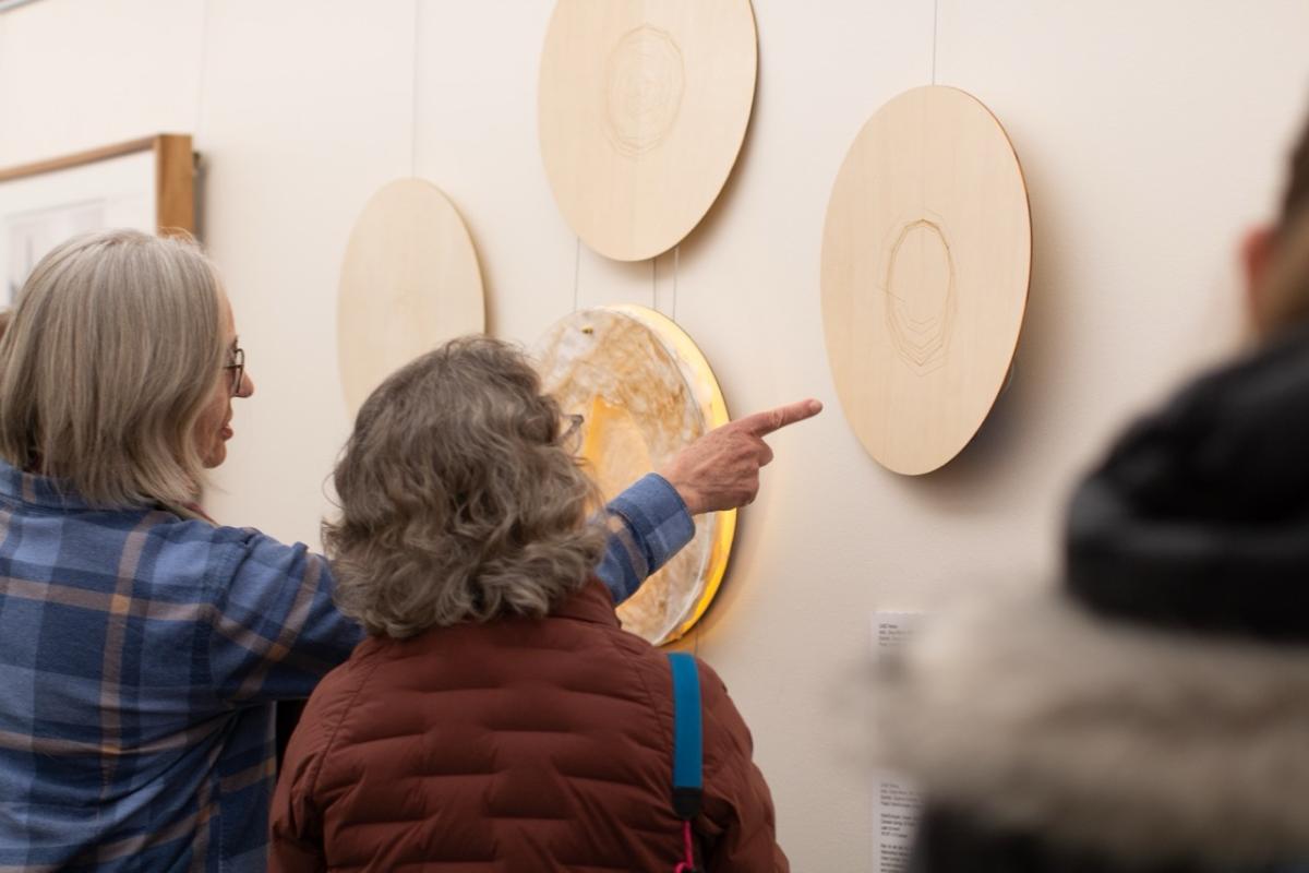 Two women stands in front of an art exhibition that features a round glowing wooden platform affixed to the wall.