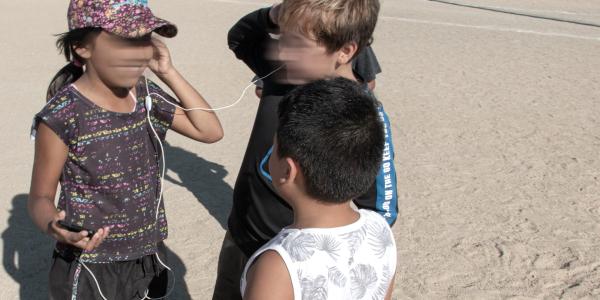 three children stand in a circle around each other. two of the children are sharing a pair of wired earbuds