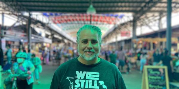 Khalil stands in front of a crowded market wearing a shirt that reads "We Still Here"