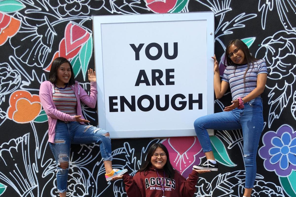 CUUB students posing in front of a mural with a sign on it that reads "you are enough"