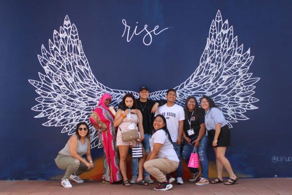 A group of students standing in front of a mural with dark blue background and white angel wings painted with the word "Rise" above them