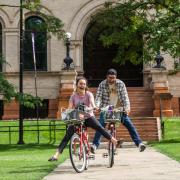 A woman and a man joyfully ride their bikes in front of a campus building.