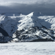 A wide shot of a snowy mountain range and cloudy skies. In the foreground, a path in the snow runs across a hill toward the mountains. Image taken from the Hindsight Journal 