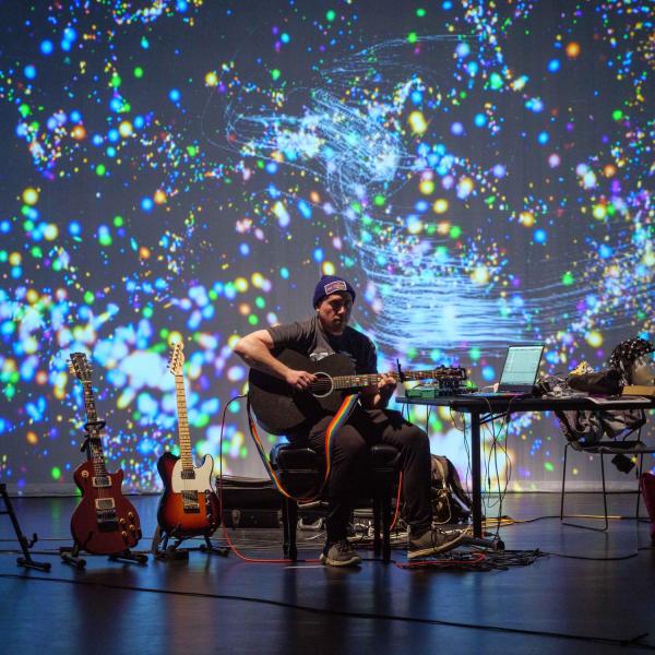 A performer play guitar in front of a projected light screen