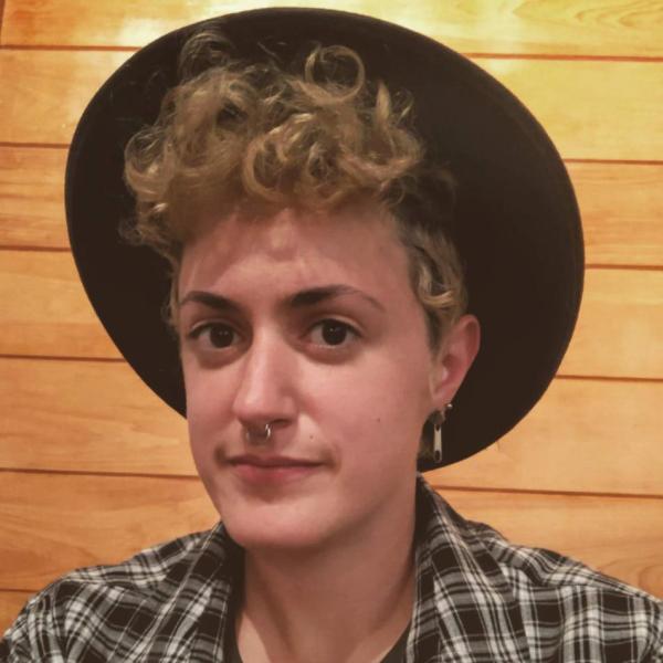A picture of Elz DeVito, a white feminine-presenting person with black and blonde curly hair covered by a black hat.