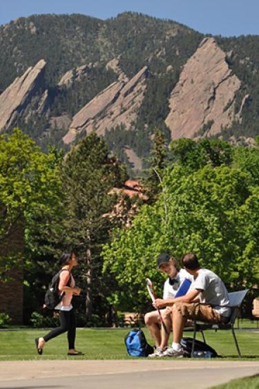 Two male students chat on a bench with a female student walks past. The Foothill mountains provide a beautiful backdrop.