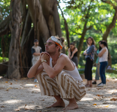 Photo of man in headband and stripped pants kneeling on ground with tree in background. 