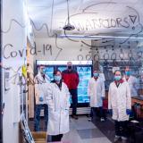 Researchers pose in their lab behind glass with the words "COVID Warriors" written on it.