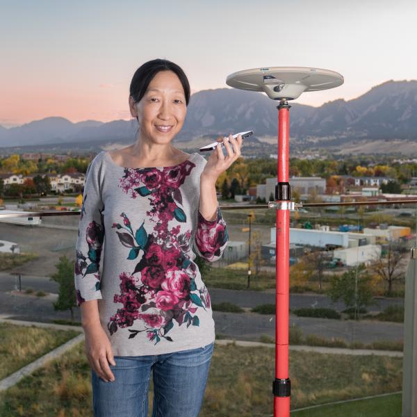 Woman standing on a balcony of a building with the Flatiron Mountains in the background. She is holding a cellphone and standing next to a small radar dish