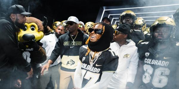 Artist Lil Wayne is surrounded by football players, Coach Prime, Chip and others. A slight mist floats in the air. (Photo by David Marckel/University of Colorado)