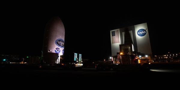 Satellite labeled "GOES" sits next to a large hangar labeled "NASA" at night time