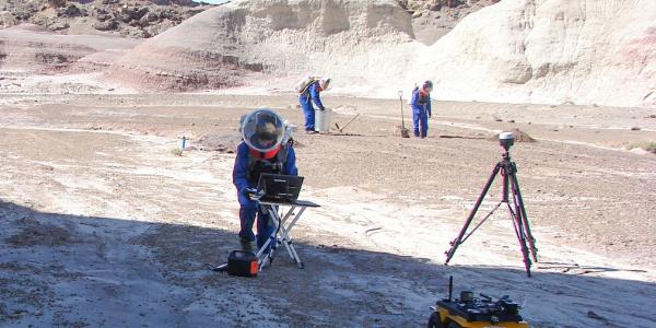 Three people in spacesuit-like gear appear to work with equipment in a dry hilly outdoor space.