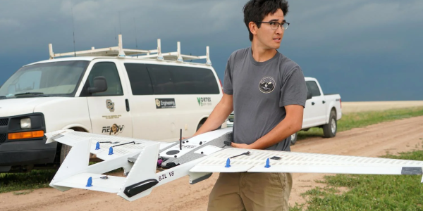 Man holds a drone with fixed wings in his arms with a white van in the background