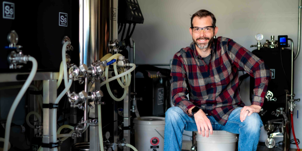 Man in flannel shirt wearing safety glasses sits next to equipment with lots of tubes