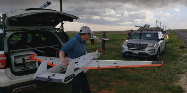 Man lifts drone with fixed wings out of the trunk of an SUV while other scientists work in background, and storm clouds brew above