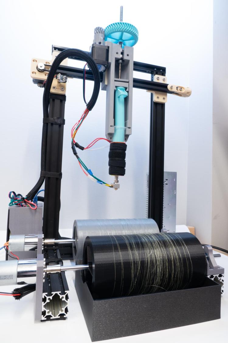 Machine sits on a tabletop with a long, plastic syringe mounted above two spinning wheels. This DIY machine for spinning gelatin fibers cost just $560 to build