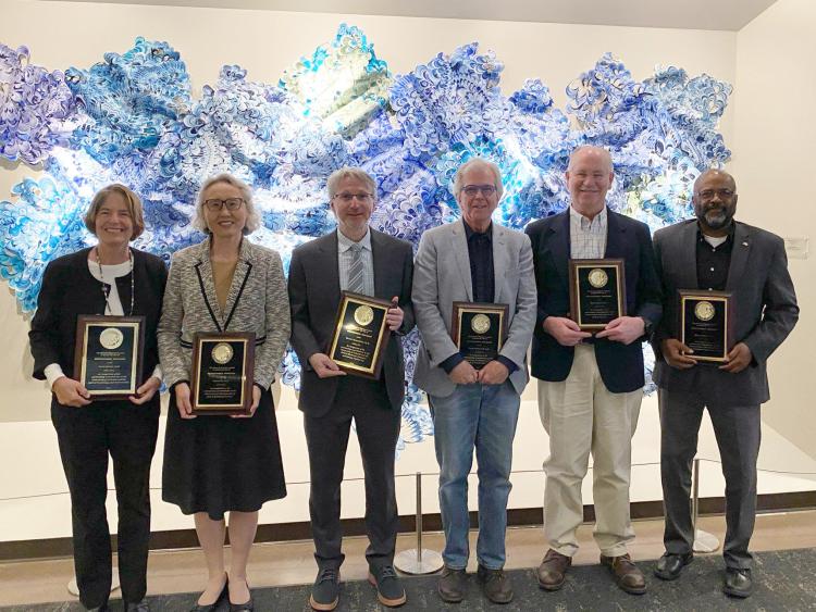 The six newest CU distinguished professors stand in a line, each holding a recognition plaque, with a large purple and blue texturized wall sculpture behind the group.