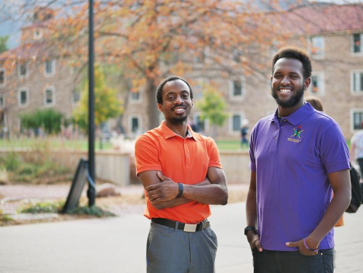 Yohannese Gebremedhin, right,  and Arthur Antoine, left, stand in front of a tree and sandstone buildings in a courtyard on the University of Colorado campus.
