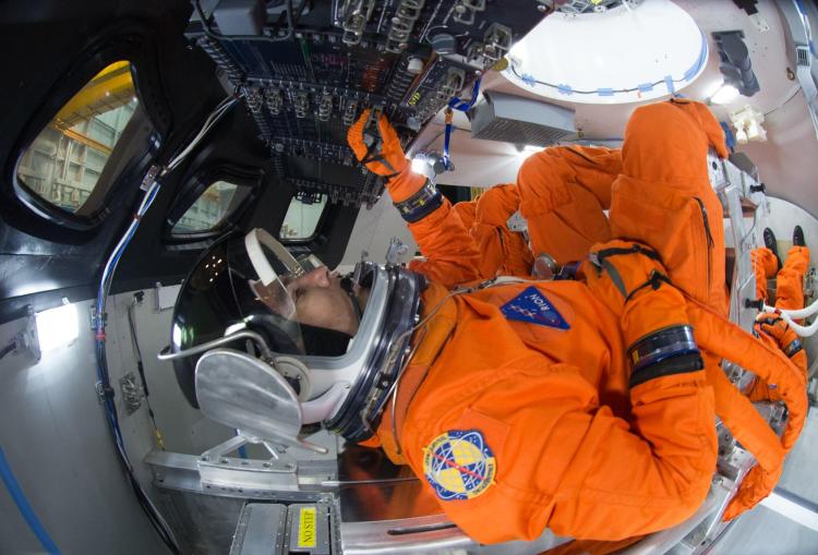 People in orange suits and helmets sit inside the cramped cockpit of a spacecraft