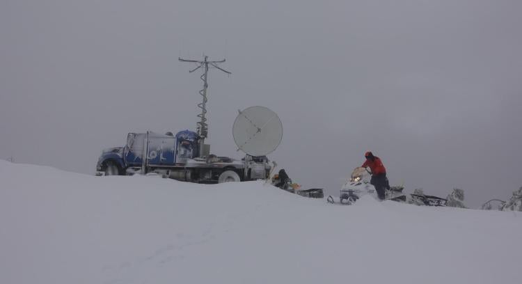 A man rides a snowmobile next to a radar dish mounted on the back of a truck.