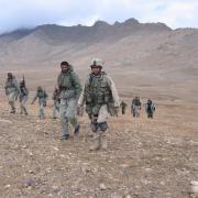 Mitch Utterback along with several other members of armed forces walk in what appears to be a dry climate, out in open space with mountains in the background.