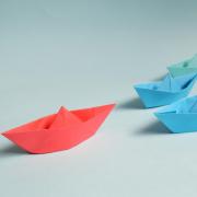 red paper boat leads a pack of blue and green paper boats