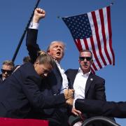 Donald Trump with an extended arm and agents surrounding him, stands on a stage, American flag draped from above, stands with streams of blood across his face.