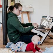 parent working on laptop while baby crawls on floor
