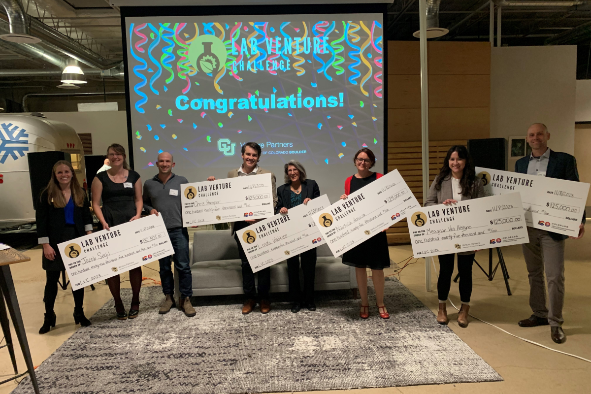 A diverse group of people stands on a presentation stage holding giant prize checks.