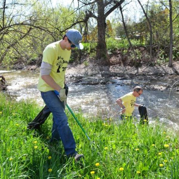 a volunteer wearing a yellow shirt with trash grabbers in right hand and trash bag in left hand near a stream on a grassy area