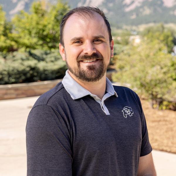 Andrew posing in front of the flatirons with a gray shirt with a white collar and small cu symbol on the right